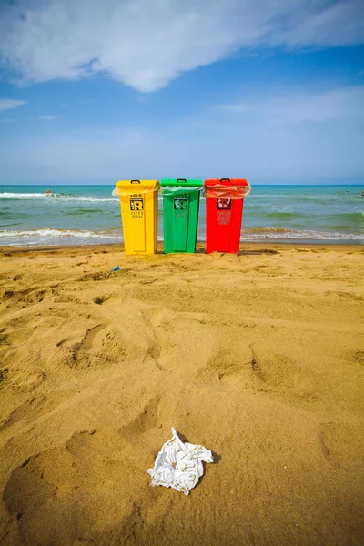 A view of three trash bin in yellow, green and red colours on a beach with some beach umbrellas