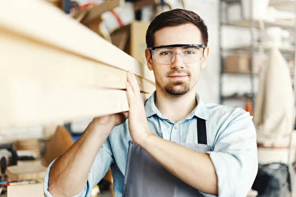 Portrait of professional young carpenter carrying wood planks on his shoulder. Young woodworker looking at camera through protective eyeglasses