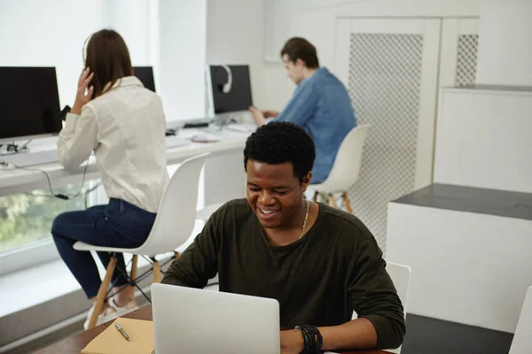 Two faceless students and young cheerful black male using laptop in modern library