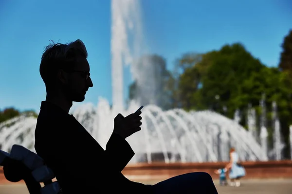 Side view of dark male figure sitting on park bench and looking at mobile phone with bright sunlit fountain on blurred background at summer