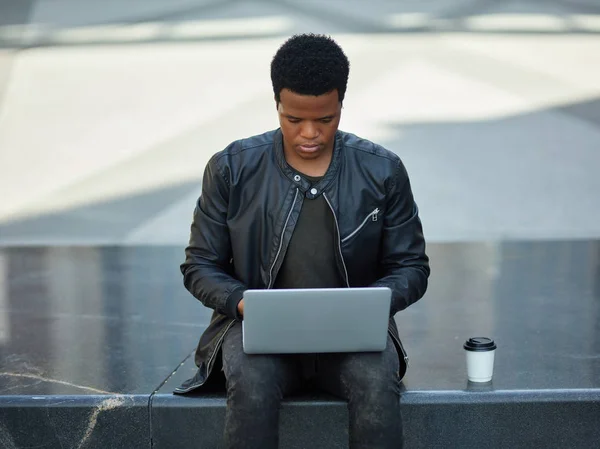 Portrait of young African man in leather jacket working on laptop outdoors, paper coffee cup o stone seat near him