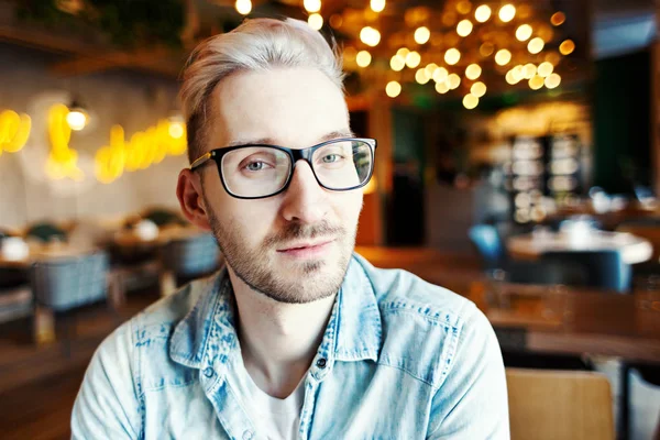 Blonde hipster man with stubble beard in eyeglasses looking at camera in cafe