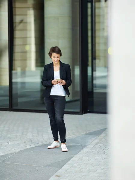 Adult elegant executive woman in black jacket and jeans standing on pavement and surfing smartphone