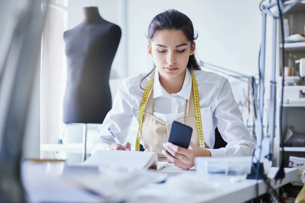 Attractive young woman with measure tape browsing smartphone and writing in notebook while sitting at table in tailor workshop