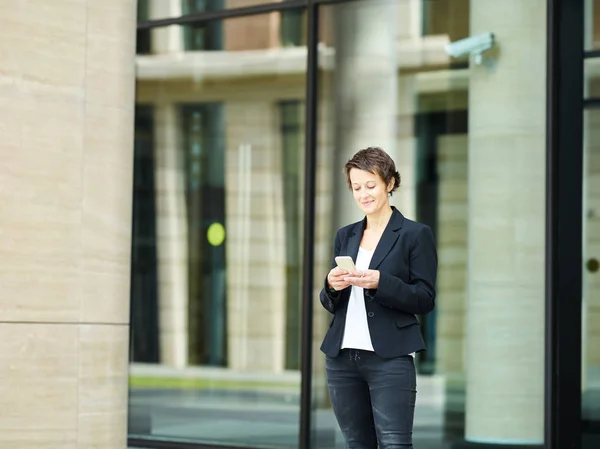 Trendy middle-aged woman in black jacket browsing smartphone with smile on background of contemporary building exterior