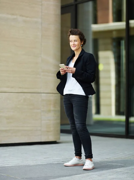 Modern middle-aged executive woman in black jacket standing on street with smartphone looking away