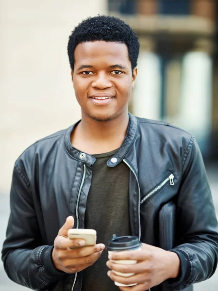 Portrait of African young man in leather jacket smiling at camera while using mobile application on his smartphone, disposable coffe cup in other hand