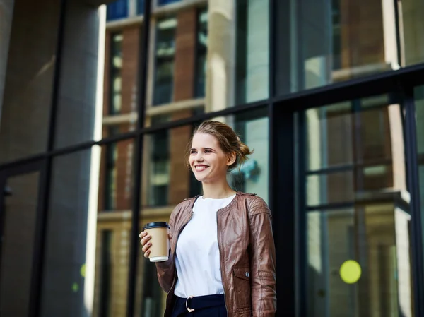 Beautiful young blond woman in jacket holding coffee cup and smiling away on background of modern office building