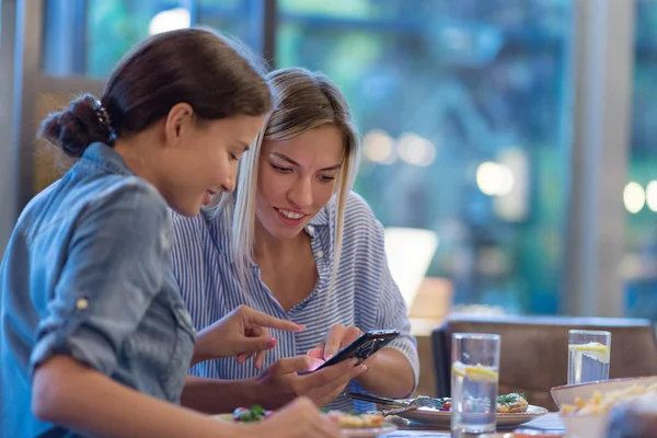 Side view of two smiling female friends browsing pictures on smart phone while having dinner at table in restaurant