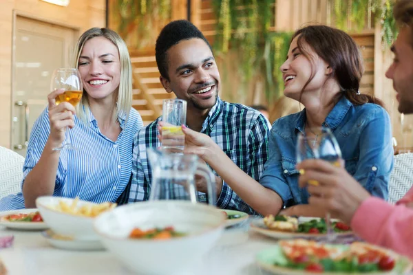 Group of four young people enjoying eating tasty food, drinking alcohol and talking at dinner in restaurant