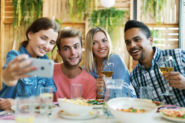 Group of four young people gathering for dinner in restaurant and posing together for selfie on smartphone