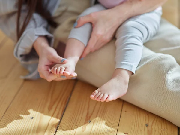 Hands of loving mother massaging cute foot of adorable baby daughter sitting on her lap