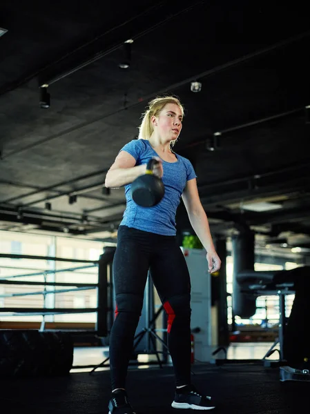 Full length three quarters portrait of strong fit woman doing weightlifting exercise with kettlebell in gym