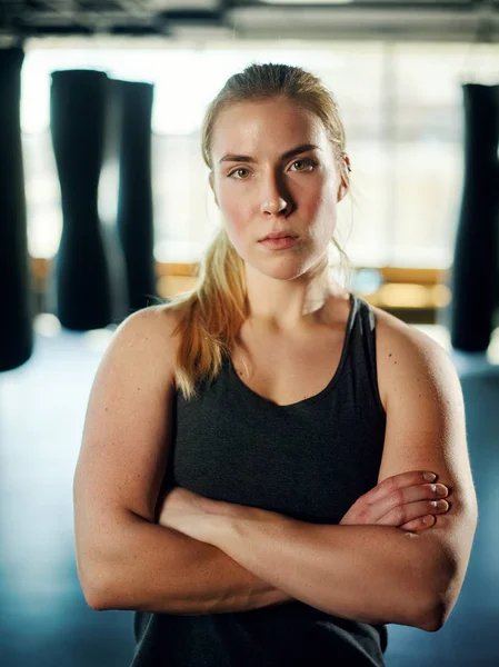Waist up portrait of strong confident woman standing with her arms crossed among boxing bags in gym