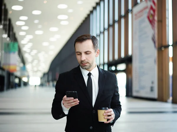 Confident middle aged manager in suit and necktie typing messages on cell phone and drinking takeaway coffee while walking along hall in business center, mall or airport