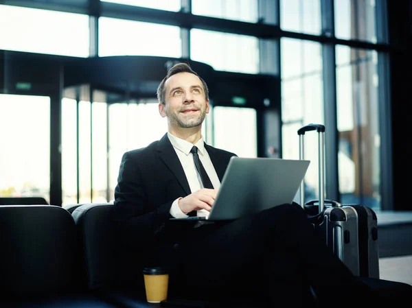 Smiling middle aged businessman sitting on leather sofa in airport lounge and looking at departure board, laptop computer on his lap and trolley suitcase placed nearby