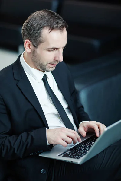 Side view of intelligent middle aged businessman in suit sitting on leather couch in airport lounge or office lobby and typing on laptop computer