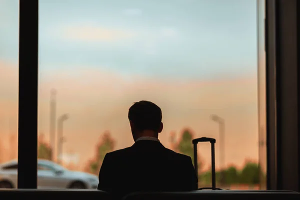 Backlit rear view of man sitting on bench in airport waiting for flight and reading book or using cell phone on background of sunset sky in window