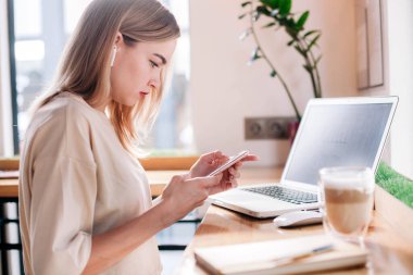 Elinde telefonla bir kafede dizüstü bilgisayarın arkasında çalışan ve cappuccino içen genç bir kız.