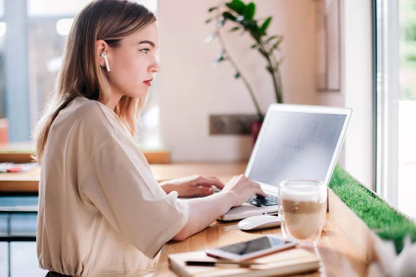 Młoda dziewczyna pracuje w laptopie w kawiarni i pije cappuccino. — Zdjęcie stockowe