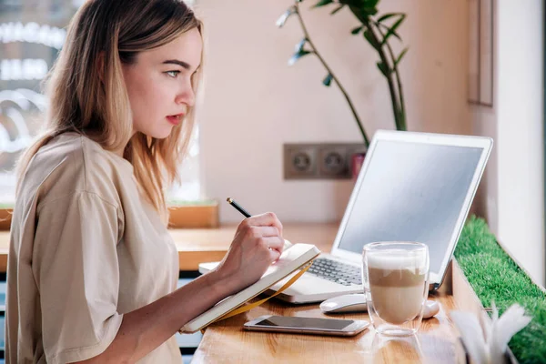 Młoda dziewczyna pracuje w laptopie w kawiarni i pije cappuccino. — Zdjęcie stockowe