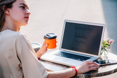 Yazın oyun bahçesindeki bir kafede laptopun arkasında çalışan ve tek kullanımlık bir bardaktan kahve içen genç bir kız. 