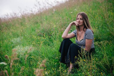 Siyah uzun etekli ve çizgili tişörtlü güzel kız yeşil bir çayırın yamacında oturuyor.