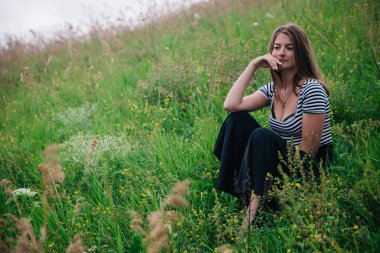 Siyah uzun etekli ve çizgili tişörtlü güzel kız yeşil bir çayırın yamacında oturuyor.