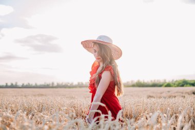 Kırmızı elbiseli ve çizgili şapkalı güzel kız buğday tarlasında poz veriyor.