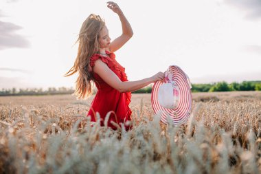 Kırmızı elbiseli ve çizgili şapkalı güzel kız buğday tarlasında poz veriyor.
