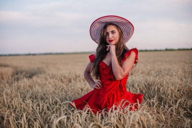 Kırmızı elbiseli ve çizgili şapkalı güzel kız buğday tarlasında poz veriyor.