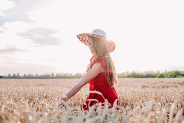 Vacker flicka i röd klänning och randig hatt poserar i ett vetefält — Stockfoto