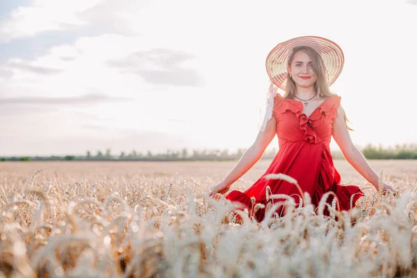 Vacker flicka i röd klänning och randig hatt poserar i ett vetefält — Stockfoto