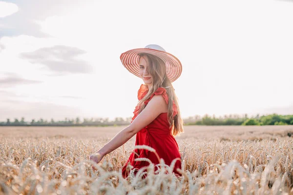 Vacker flicka i röd klänning och randig hatt poserar i ett vetefält — Stockfoto