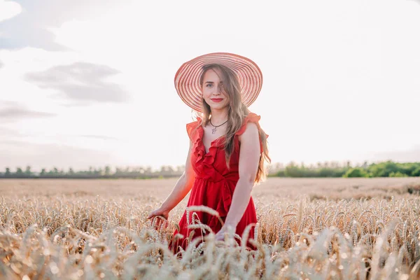 Vacker flicka i röd klänning och randig hatt poserar i ett vetefält — Stockfoto