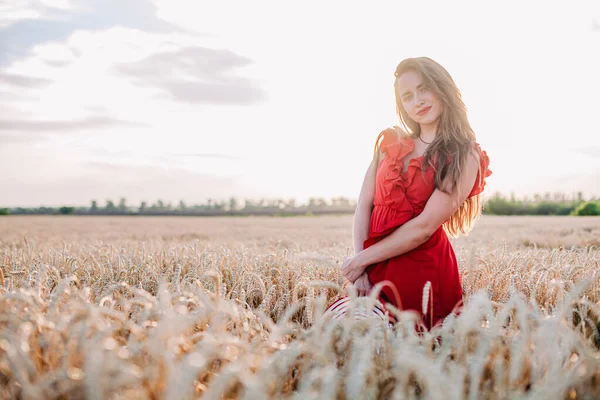 Vacker flicka i röd klänning och randig hatt poserar i ett vetefält — Stockfoto