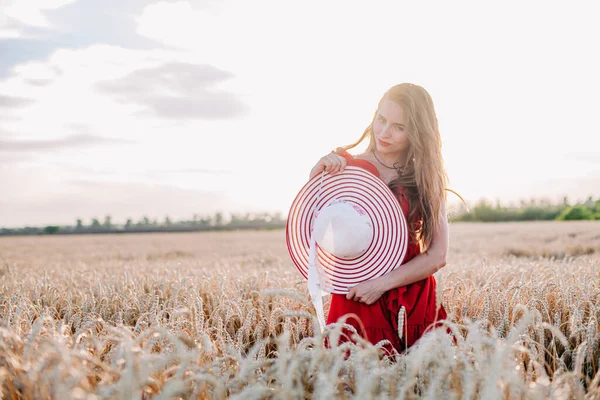 Vacker flicka i röd klänning och randig hatt poserar i ett vetefält — Stockfoto