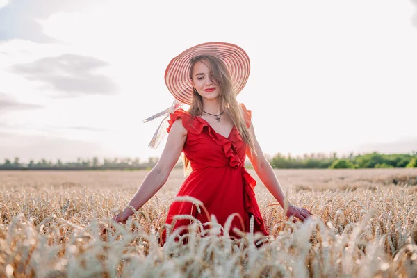 Kırmızı elbiseli ve çizgili şapkalı güzel kız buğday tarlasında poz veriyor. — Stok fotoğraf