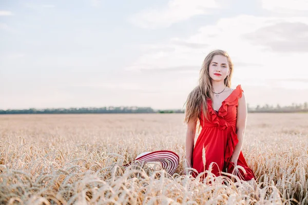Vacker flicka i röd klänning och randig hatt poserar i ett vetefält — Stockfoto