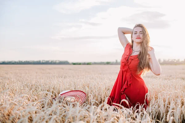 Vacker flicka i röd klänning och randig hatt poserar i ett vetefält — Stockfoto