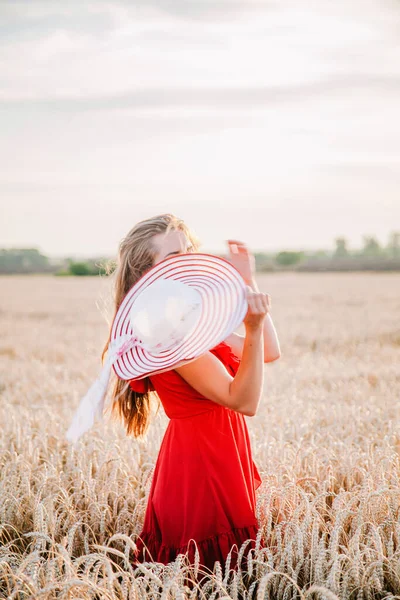 Vacker flicka i röd klänning och randig hatt poserar i ett vetefält — Stockfoto