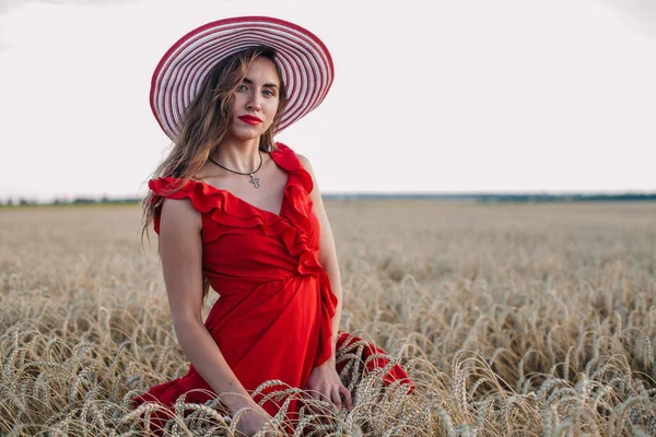 Menina bonita em vestido vermelho e chapéu listrado posando em um campo de trigo — Fotografia de Stock