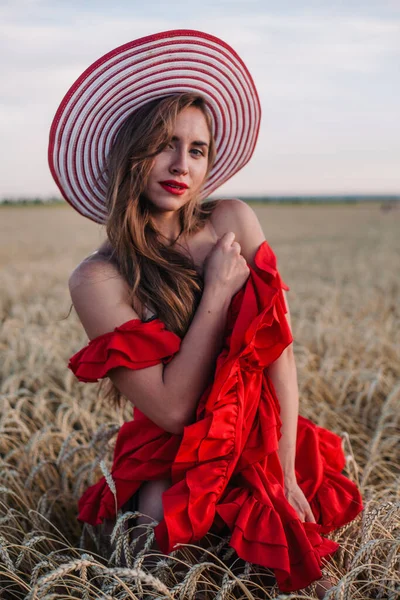 Krásná dívka v červených šatech a pruhované klobouk pózování v pšeničném poli — Stock fotografie