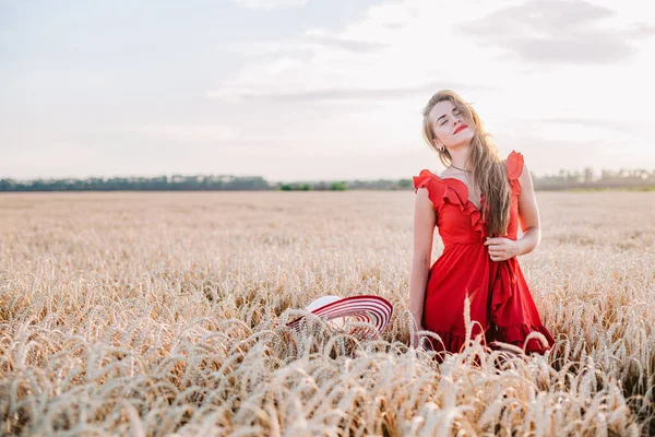 Όμορφο κορίτσι με κόκκινο φόρεμα και ριγέ καπέλο ποζάροντας σε χωράφι με σιτάρι — Φωτογραφία Αρχείου