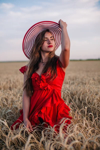 Menina bonita em vestido vermelho e chapéu listrado posando em um campo de trigo — Fotografia de Stock