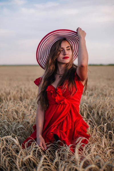 Piękna dziewczyna w czerwonej sukience i kapelusz w paski pozowanie na polu pszenicy — Zdjęcie stockowe