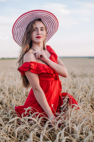 Kırmızı elbiseli ve çizgili şapkalı güzel kız buğday tarlasında poz veriyor. — Stok fotoğraf