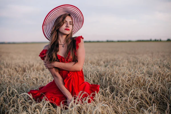 Menina bonita em vestido vermelho e chapéu listrado posando em um campo de trigo — Fotografia de Stock