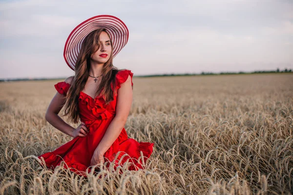 Vacker flicka i röd klänning och randig hatt poserar i ett vetefält — Stockfoto