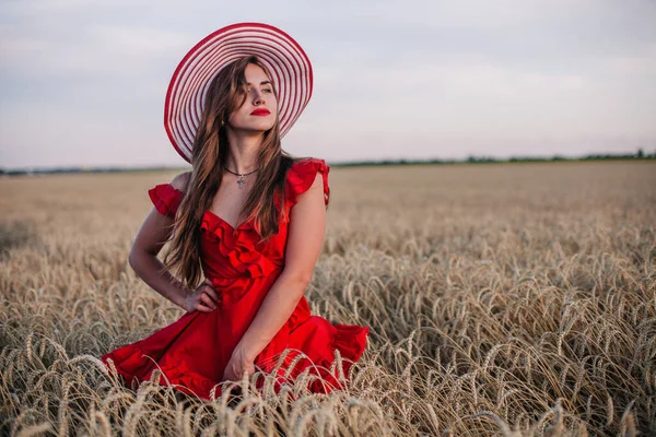 Menina bonita em vestido vermelho e chapéu listrado posando em um campo de trigo — Fotografia de Stock